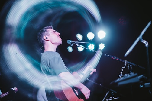 A musician passionately singing and playing an acoustic guitar on stage. There's a dynamic motion effect with swirling lights around the performer, while bright spotlights shine in the background.