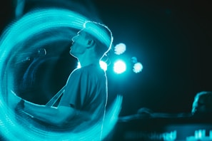 A person is playing an electric guitar with a swirl of blue light surrounding them. The background includes bright stage lights creating a glowing effect around the silhouette. The atmosphere suggests a live music performance with a dynamic and vibrant visual effect.