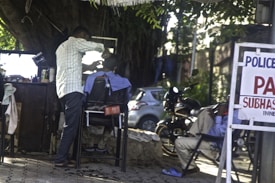 A barber is giving a haircut to a customer seated outdoors. The setting is informal, with a mirror and various haircutting tools placed on a wooden surface. Another person is sitting nearby, possibly waiting. There is a sign with text visible, along with a motorcycle and a tree providing shade.