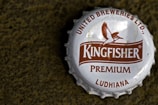 A close-up view of a metallic bottle cap with the brand name 'Kingfisher' prominently displayed in the center. The cap features a logo of a bird in flight and text indicating 'United Breweries Ltd.', 'Premium', and 'Ludhiana'. The cap is white with brown text and logo, and it rests on a rough textured dark surface.