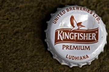A close-up view of a metallic bottle cap with the brand name 'Kingfisher' prominently displayed in the center. The cap features a logo of a bird in flight and text indicating 'United Breweries Ltd.', 'Premium', and 'Ludhiana'. The cap is white with brown text and logo, and it rests on a rough textured dark surface.