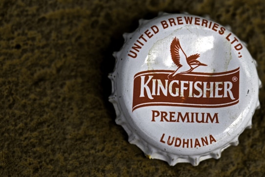 A close-up view of a metallic bottle cap with the brand name 'Kingfisher' prominently displayed in the center. The cap features a logo of a bird in flight and text indicating 'United Breweries Ltd.', 'Premium', and 'Ludhiana'. The cap is white with brown text and logo, and it rests on a rough textured dark surface.