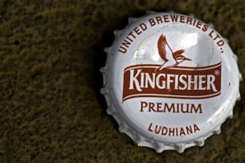 A close-up view of a metallic bottle cap with the brand name 'Kingfisher' prominently displayed in the center. The cap features a logo of a bird in flight and text indicating 'United Breweries Ltd.', 'Premium', and 'Ludhiana'. The cap is white with brown text and logo, and it rests on a rough textured dark surface.