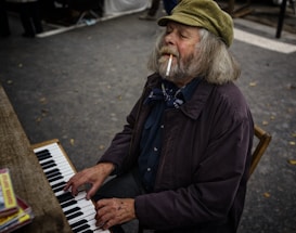 An elderly man with long hair and a beard is playing a keyboard while sitting on a wooden chair. He is wearing a brown jacket, a corduroy cap, and has a cigarette in his mouth. The scene is outdoors, possibly at a market or street event, with some scattered leaves on the ground.