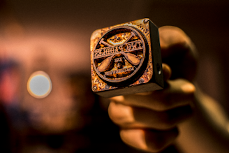 A hand holding a close-up view of a metal stamp with a rustic, worn appearance. The stamp features text and symbols, likely used for branding or decorative purposes. The background is blurred with warm tones, enhancing the focus on the stamp.