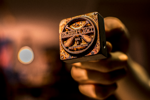 A hand holding a close-up view of a metal stamp with a rustic, worn appearance. The stamp features text and symbols, likely used for branding or decorative purposes. The background is blurred with warm tones, enhancing the focus on the stamp.