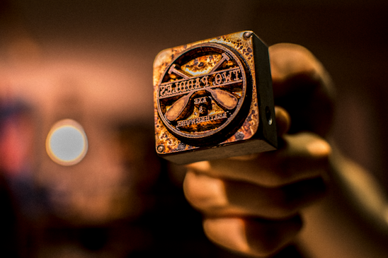 A hand holding a close-up view of a metal stamp with a rustic, worn appearance. The stamp features text and symbols, likely used for branding or decorative purposes. The background is blurred with warm tones, enhancing the focus on the stamp.