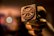A hand holding a close-up view of a metal stamp with a rustic, worn appearance. The stamp features text and symbols, likely used for branding or decorative purposes. The background is blurred with warm tones, enhancing the focus on the stamp.