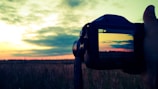 A hand holding a camera against a landscape backdrop, capturing a picturesque sunset over a field. The camera display mirrors the vibrant sky, featuring a gradient of warm colors as clouds drift at the horizon.