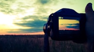 A hand holding a camera against a landscape backdrop, capturing a picturesque sunset over a field. The camera display mirrors the vibrant sky, featuring a gradient of warm colors as clouds drift at the horizon.
