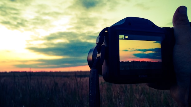A hand holding a camera against a landscape backdrop, capturing a picturesque sunset over a field. The camera display mirrors the vibrant sky, featuring a gradient of warm colors as clouds drift at the horizon.