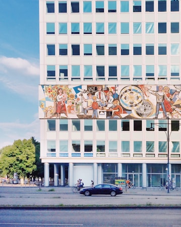 A large building facade featuring a vibrant, colorful mural depicting various figures engaged in activities such as science with a telescope, family gatherings, and labor. Below the mural, a modern multistory building with grid-like windows is visible, along with a street scene including a car and several cyclists.