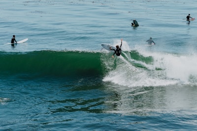 Several surfers are riding and waiting for waves in the ocean. One surfer is actively maneuvering on a wave, creating a dynamic splash, while others are positioned on their boards, either watching or paddling.