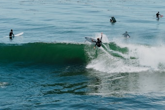 Several surfers are riding and waiting for waves in the ocean. One surfer is actively maneuvering on a wave, creating a dynamic splash, while others are positioned on their boards, either watching or paddling.