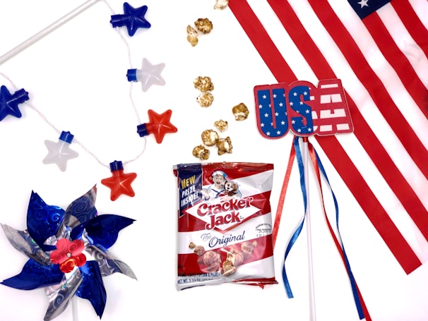 An arrangement featuring American-themed items, including a pinwheel with red, white, and blue blades, a string of star-shaped lights, a plastic card with 'USA' on it, a bag of Cracker Jack popcorn, and a section of the American flag. The festive items are spread against a white background.