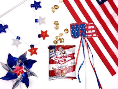 An arrangement featuring American-themed items, including a pinwheel with red, white, and blue blades, a string of star-shaped lights, a plastic card with 'USA' on it, a bag of Cracker Jack popcorn, and a section of the American flag. The festive items are spread against a white background.