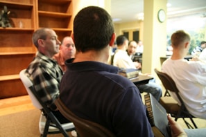 A group of people sit in a room with bookshelves and chairs. Some are engaged in conversation, while others face forward attentively. The room has a casual and informal setting, with a mix of focused and relaxed postures.