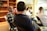 A group of people sit in a room with bookshelves and chairs. Some are engaged in conversation, while others face forward attentively. The room has a casual and informal setting, with a mix of focused and relaxed postures.