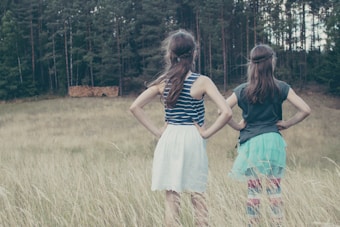 Two girls stand in a grassy field with their backs to the camera, looking towards a dense forest. One wears a striped tank top and white skirt, the other a dark top and blue skirt. The scene is set in a serene, natural environment.