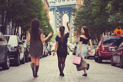 Three women are walking down a cobblestone street lined with parked cars and trees. Two women are carrying shopping bags, one of which reads 'SALE'. In the distance, the base of a large bridge is visible, framed by the buildings along the street. The scene conveys a sense of urban leisure and socializing.