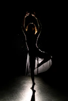 A wide shot of a dancer mid-leap, her silhouette outlined against a deep black background.