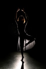 A silhouette of a dancer striking a pose, with one leg raised and arms arched above the head. The light highlights the edges of the dancer, creating a striking contrast against a dark background.