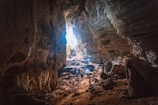 A vast cave with rugged walls and a large opening, allowing natural light to illuminate the space. Several people are standing inside, some holding lights or cameras, suggesting exploration or photography. The cave's interior is rugged and natural, with rocks scattered across the ground.