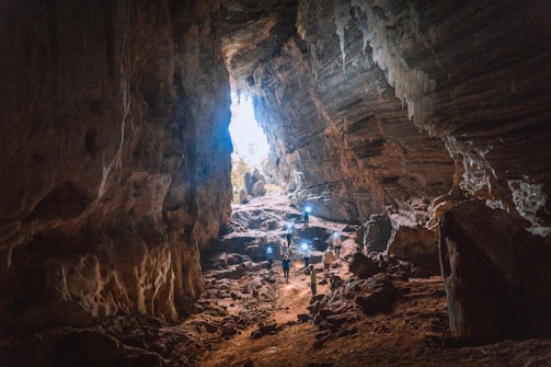 A vast cave with rugged walls and a large opening, allowing natural light to illuminate the space. Several people are standing inside, some holding lights or cameras, suggesting exploration or photography. The cave's interior is rugged and natural, with rocks scattered across the ground.