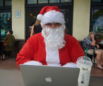 A person dressed as Santa Claus is seated at a table outdoors, using a laptop with a visible Apple logo. In front of them is a cold beverage from Starbucks with a green straw. The background shows a caf&eacute; setting with other people seated and engaged in conversations.
