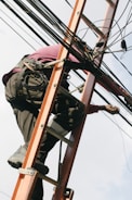 A person is climbing a ladder while working on electrical or communication cables. They are wearing work boots and protective gear, including knee pads, and have a harness for safety. Various cables are intertwined around the ladder.