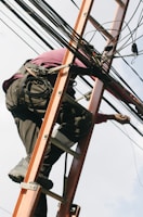 A person is climbing a ladder while working on electrical or communication cables. They are wearing work boots and protective gear, including knee pads, and have a harness for safety. Various cables are intertwined around the ladder.
