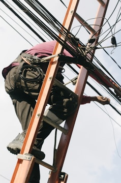 A person is climbing a ladder while working on electrical or communication cables. They are wearing work boots and protective gear, including knee pads, and have a harness for safety. Various cables are intertwined around the ladder.