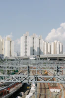 Numerous tall, white residential buildings stand against a clear sky. Below, multiple train tracks intersect with overhead power lines. There is a train moving along the bottom track, and a construction vehicle is visible nearby. The scene conveys an urban setting with infrastructure and residential areas coexisting.