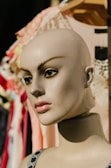A mannequin head with a smooth, painted complexion and detailed facial features, including defined eyebrows and large, expressive eyes. It is situated against a backdrop of various colorful fabrics and textiles that hang on a rack.