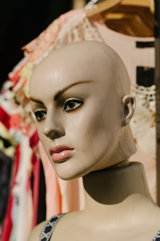 A mannequin head with a smooth, painted complexion and detailed facial features, including defined eyebrows and large, expressive eyes. It is situated against a backdrop of various colorful fabrics and textiles that hang on a rack.