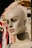 A mannequin head with a smooth, painted complexion and detailed facial features, including defined eyebrows and large, expressive eyes. It is situated against a backdrop of various colorful fabrics and textiles that hang on a rack.