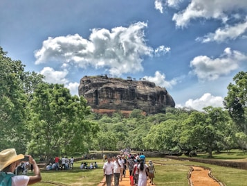 A large rock formation rises majestically in the background, surrounded by lush green vegetation and trees under a partly cloudy sky. In the foreground, people are walking on a dirt path, with some wearing hats and taking photos, enjoying a day out in nature.