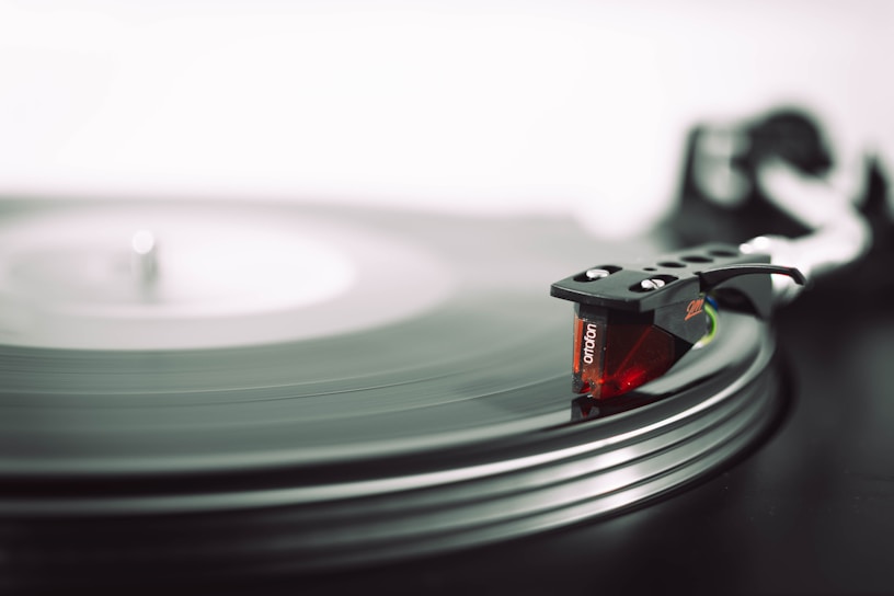 A close-up of a vinyl record being played on a turntable with a red and black cartridge arm. The record is spinning and captures the groove lines in detail.
