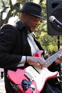A person wearing a fedora and coat plays an electric guitar. The red guitar has a white pickguard and is connected to an amplifier. The individual appears focused on playing music, standing outdoors with trees visible in the background.