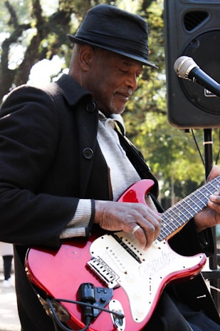 A person wearing a fedora and coat plays an electric guitar. The red guitar has a white pickguard and is connected to an amplifier. The individual appears focused on playing music, standing outdoors with trees visible in the background.