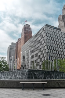 A cityscape with modern high-rise buildings, one of which has the logo of a well-known bank. A large, tiered fountain is in the foreground, with water cascading down its steps. The background features a tall building topped with an American flag.