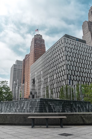 A cityscape with modern high-rise buildings, one of which has the logo of a well-known bank. A large, tiered fountain is in the foreground, with water cascading down its steps. The background features a tall building topped with an American flag.