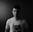 A monochrome portrait of a young man standing against a dark wooden background. He is wearing a T-shirt with a graphic print of a masked character, possibly a superhero. The lighting creates a dramatic contrast with shadows covering one side of his face.
