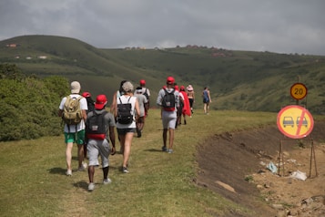 A group of people walking along a grassy path in a hilly area. They wear casual outdoor clothing, and several have backpacks. Red caps are a common accessory among the group. Road signs indicating a speed limit and no entry for cars are present on the right side.