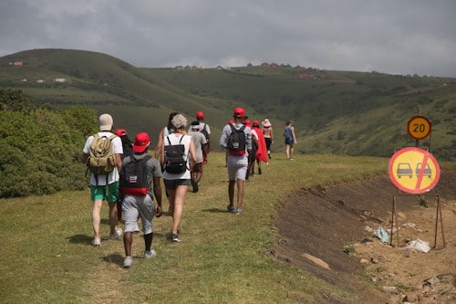 A group of people walking along a grassy path in a hilly area. They wear casual outdoor clothing, and several have backpacks. Red caps are a common accessory among the group. Road signs indicating a speed limit and no entry for cars are present on the right side.
