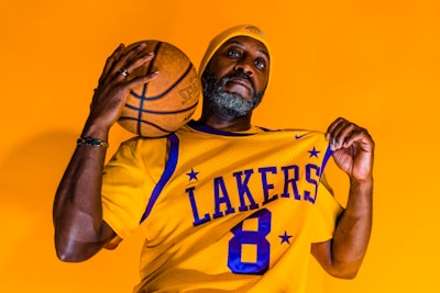 A person wearing a yellow basketball jersey with 'LAKERS' and the number 8, holding a basketball and a matching beanie. The background is a solid bright orange, highlighting the vibrant colors of both the attire and the backdrop.