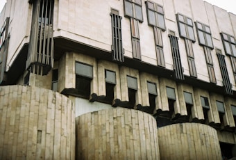 A brutalist-style architectural building featuring large concrete pillars and protruding rectangular windows. The structure has a rugged, geometric appearance with vertical lines and a muted color palette.