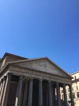 A classical Roman building with a grand portico and large Corinthian columns. The triangular pediment features an ancient inscription. The structure is set against a clear blue sky, and part of a modern building is visible in the background.