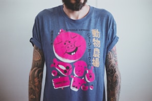 A person wearing a blue T-shirt with rolled-up sleeves, featuring a large pink graphic of a cartoon pig and Japanese text. The shirt references George Orwell's 'Animal Farm'. The person's arms have tattoos, and the background is a plain light color.
