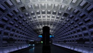 A dimly lit underground metro station with an arched, geometric ceiling design. A train is approaching on the track with bright headlights illuminating the dark surroundings. A digital display board is visible in the distance showing train information. The overall architecture features repetitive patterns and a modern, industrial aesthetic.